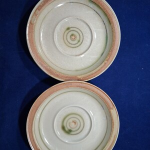 May include: Two ceramic plates with a cream-colored center and a concentric spiral design. The plates have a light brown rim and a green accent ring. The plates are on a dark blue surface.