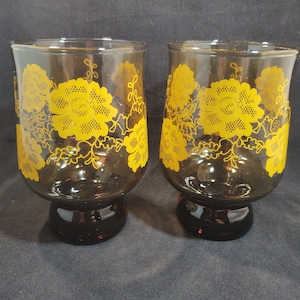 May include: Two amber glass drinking glasses with yellow floral designs. The glasses have a flared base.