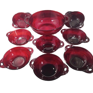 May include: A collection of ruby red glass bowls, comprising a large serving bowl and smaller bowls with handles. The bowls feature a textured design and are arranged on a white backdrop. The set is likely for serving food.