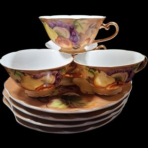 May include: A set of three porcelain teacups and matching saucers featuring a hand-painted fruit motif with pears, grapes, and peaches. The pieces have a warm tan background, scalloped edges, and gold-toned trim on the handles and rims.