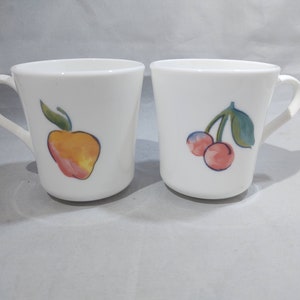 May include: Two white ceramic mugs with handles. One mug has a hand-painted image of a red apple with green leaves. The other mug has a hand-painted image of two red cherries with green leaves.