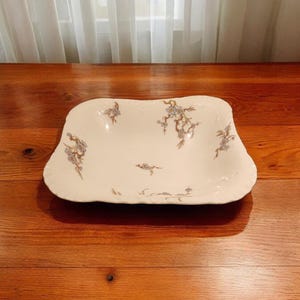 May include: A white square serving bowl with a floral design featuring blue flowers and brown stems. The bowl has a slightly scalloped edge.