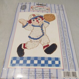 Może przedstawiać: Zestaw do haftu krzyżykowego z Raggedy Ann & Andy. Projekt przedstawia wyszytą krzyżykami postać Raggedy Ann w stroju baseballowym, trzymającą rękawicę. Tło ma niebiesko-biały wzór w kratkę i pasiastą obwódkę.