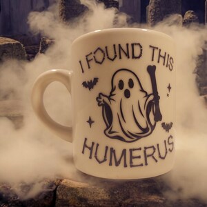 May include: A white ceramic mug with a black ghost holding a bone and the text "I found this humerus" on it. The mug is sitting on a rock with smoke in the background.