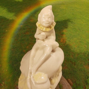 May include: A white ceramic figurine of a leprechaun sitting on a pot of gold. The leprechaun is wearing a yellow hat and a green vest. The figurine is sitting on a rock with a rainbow in the background.