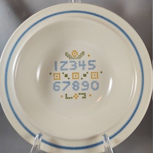May include: White ceramic bowl with a blue rim and a cross-stitch design of numbers 12345, 67890, and L7. The bowl is on a white stand.