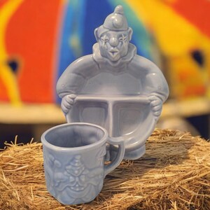 May include: A blue ceramic clown-shaped divided plate and a matching blue ceramic cup with a clown design. The plate has three sections and the cup has a handle.