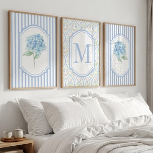 May include: A set of three framed wall art prints featuring light blue vertical stripes. The outer prints display blue hydrangea illustrations within decorative borders, while the center print features a large letter M on a floral pattern.