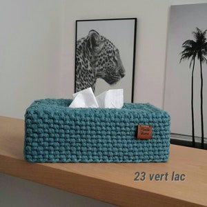 Op de afbeelding: Een rechthoekige, tealgekleurde gehaakte tissueboxhoes met een gestructureerd, geweven steekpatroon. Een klein bruin leren label met de tekst "Maille Home" is aan de zijkant bevestigd. Twee witte tissues steken uit de opening aan de bovenkant.