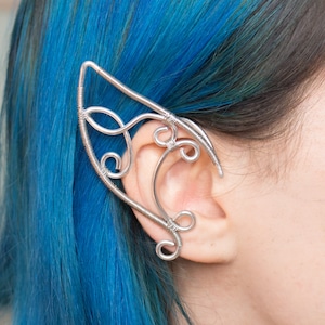 Może przedstawiać: Srebrny kolczyk na ucho w kształcie mankietu o delikatnym, skomplikowanym wzorze. Mankiet jest noszony na górnej części ucha, a wzór rozciąga się wokół płatka ucha.