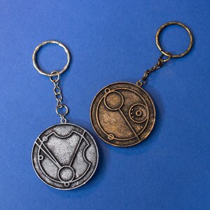 May include: Two silver and bronze keychains with circular pendants featuring intricate designs. The pendants are reminiscent of a clock face with Roman numerals and other markings.