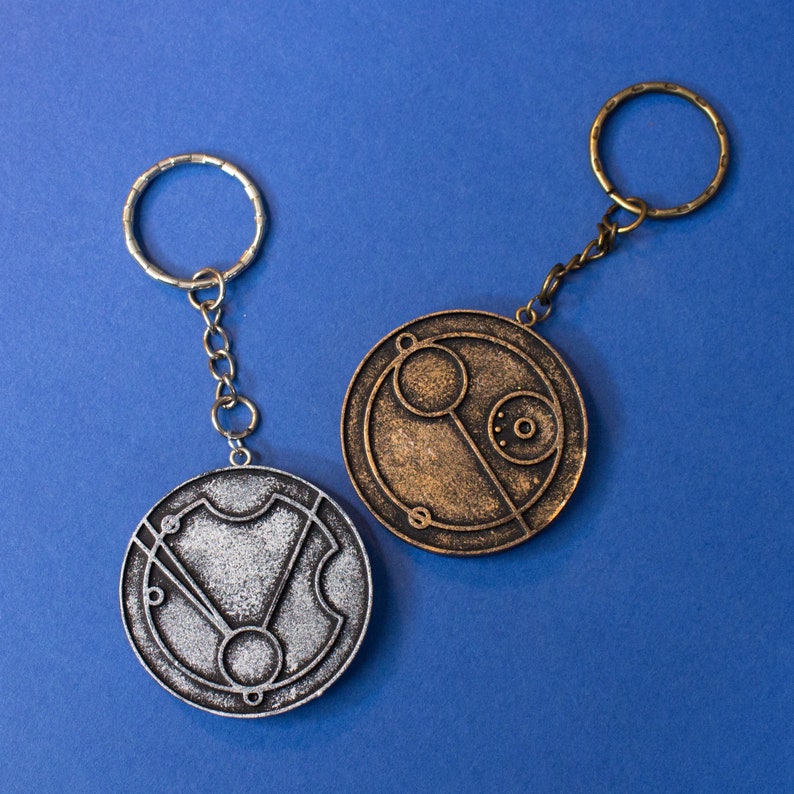 May include: Two silver and bronze keychains with circular pendants featuring intricate designs. The pendants are reminiscent of a clock face with Roman numerals and other markings.