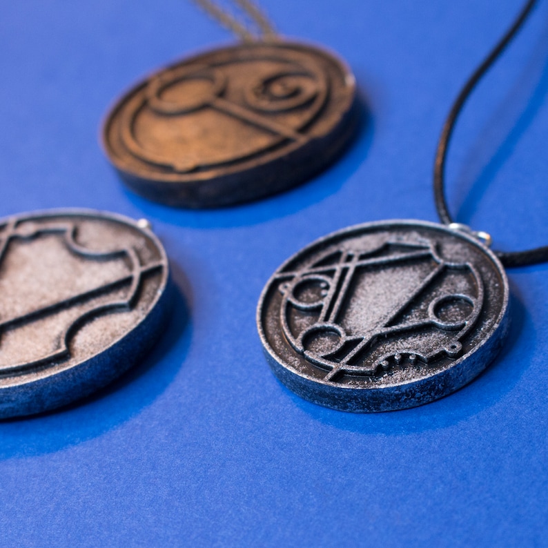 May include: Three silver and bronze circular pendants with intricate designs on a blue background. The pendants are on a black cord and have a worn, aged look.
