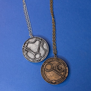 May include: Two silver and gold necklaces with circular pendants featuring intricate designs. The pendants are reminiscent of a clock face with Roman numerals and a compass.