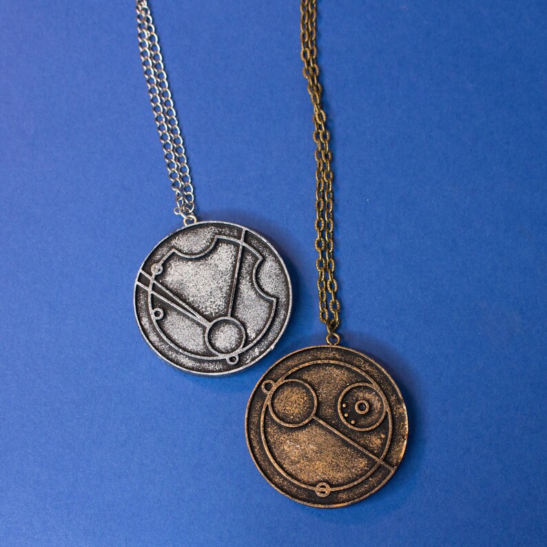 May include: Two silver and gold necklaces with circular pendants featuring intricate designs. The pendants are reminiscent of a clock face with Roman numerals and a compass.