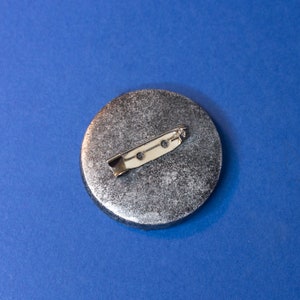 May include: A silver round pinback button with a textured surface and a silver metal pin on the back.