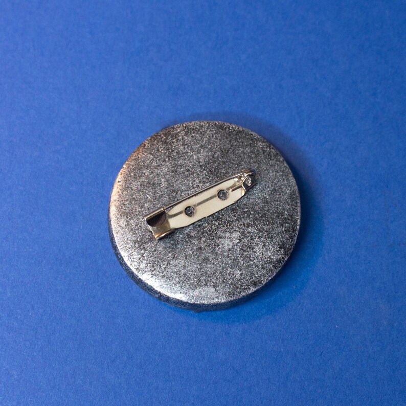 May include: A silver round pinback button with a textured surface and a silver metal pin on the back.