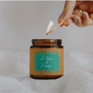 May include: A brown glass jar candle with a label that reads "Lotus & Lime" in white script. A lit match is held above the candle, with a flame. The candle sits on a white, textured surface.