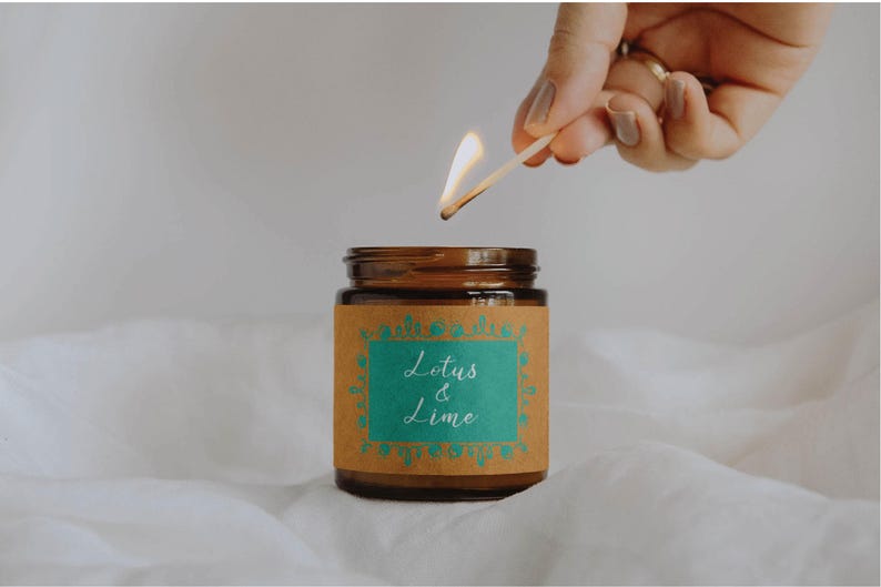 May include: A brown glass jar candle with a label that reads "Lotus & Lime" in white script. A lit match is held above the candle, with a flame. The candle sits on a white, textured surface.