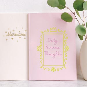 May include: Two notebooks, one light pink and one cream, are displayed. The pink notebook has a yellow frame with the text "Only Princess Thoughts." The cream notebook has gold text and stars. A white vase holds a green plant.