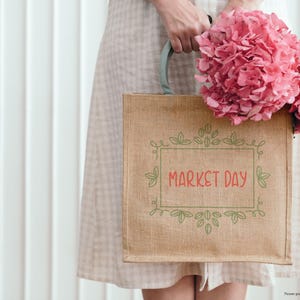 May include: A tan burlap tote bag with the words "MARKET DAY" in red script, framed by green leaves and vines. The bag is held by a person wearing a light-colored dress and holding a bouquet of pink flowers.