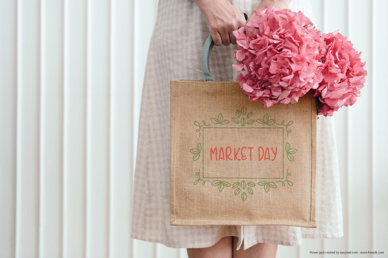 May include: A tan burlap tote bag with the words "MARKET DAY" in red script, framed by green leaves and vines. The bag is held by a person wearing a light-colored dress and holding a bouquet of pink flowers.