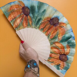 May include: A folding hand fan featuring a watercolor painting of three orange coneflowers with dark brown centers against a green and blue background. The fan has a light-colored wooden frame with visible ribs and a pleated fabric surface.
