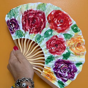 May include: A hand fan with a watercolor floral design. The fan features red, purple, and orange roses with green leaves on a white background. The fan's wooden frame is visible at the bottom.