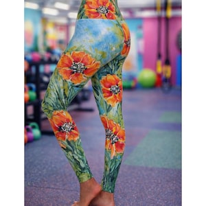 May include: High-waisted leggings featuring a vibrant watercolor-style print of large orange poppy flowers with green stems and leaves against a light blue background. The fitted garment extends to the ankles and displays a repeating floral pattern.