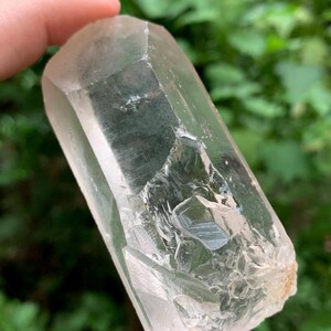 May include: A clear quartz crystal with a natural, rough base and a polished, faceted top. The crystal is held against a blurred green background, showcasing its translucent quality and internal structures. The crystal is approximately 10 cm long.