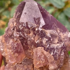 May include: A raw amethyst crystal cluster. The main point is a deep purple colour, with a faceted surface. The base is rough and textured, with varying shades of purple and light brown. The crystal has a natural, unpolished appearance.