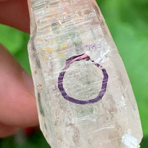 Op de afbeelding: Een heldere kwartskristal met een paarse cirkel erop getekend. Het kristal heeft een lichte, doorschijnende kleur met zichtbare interne lijnen en een enigszins ruwe textuur. De achtergrond is een vage groene kleur.