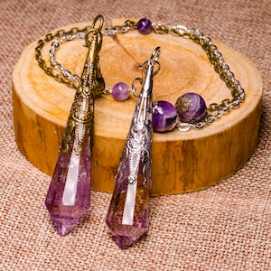 Pode incluir: Dois pêndulos de cristal de ametista com detalhes ornamentados em metal prateado e dourado pendurados em uma corrente prateada sobre uma superfície de madeira.