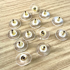 Pode incluir: Um close-up de um grupo de 13 costas de brincos de plástico transparente com postes de metal dourado. As costas são projetadas para prender os brincos no lugar.