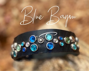 May include: A black leather bracelet with a row of blue, green, and clear rhinestones. The bracelet is decorated with silver accents and the text "Blue Bayou" is written above it.
