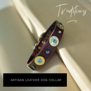Op de afbeelding: Een bruine leren hondenriem met gouden schotpatronen en blauwe strasssteentjes. De riem ligt op een beige oppervlak en de tekst "Traditions" is in cursief boven de riem geschreven.