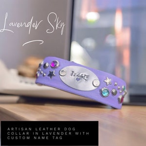 May include: A lavender leather dog collar with a custom name tag. The collar is adorned with various gemstones and star-shaped embellishments. The name "Tillie" is engraved on the silver name tag. The text "Lavender Sky" is written in cursive.
