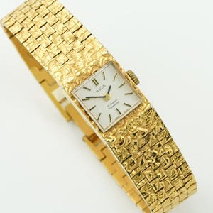 May include: A gold-toned Avia wristwatch with a textured band and square face. The watch face is white with black hands and markers. The band has a woven pattern. The watch is displayed on a white background.