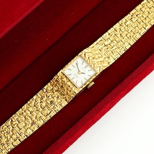 May include: A gold-toned wristwatch with a rectangular face and a textured band, resting in a red velvet box. The watch face is white with black hands and markers. The band has a woven pattern.