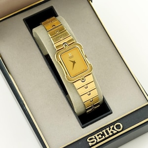 May include: A gold-toned Seiko watch with a rectangular face and matching band, presented in a black box with gold lettering. The watch is displayed on a beige cushion inside the box, which is open.