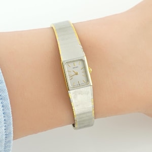 May include: A Seiko wristwatch featuring a rectangular silver-toned dial with gold-toned hands and hour markers. The watch has a two-tone band with a brushed silver centre and gold-toned edges, secured by a metal clasp on the side of the case.