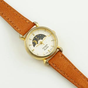 Peut inclure: Une montre Imagio Quartz dorée avec un bracelet en cuir marron. Le cadran de la montre présente un motif de lune et d'étoiles. Le bracelet mesure environ 2 cm de large.