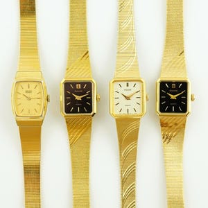 Puede incluir: Cuatro relojes de pulsera de tono dorado con diferentes caras de colores. Los relojes tienen una cara cuadrada y una banda de metal de tono dorado. Las caras son blancas, negras y crema.