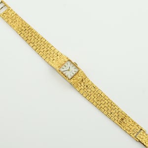 May include: A gold-toned wristwatch with a textured band and rectangular face. The watch features a white dial with black hands and markers. The band is made of small, interconnected textured gold links. The watch is displayed against a white background.