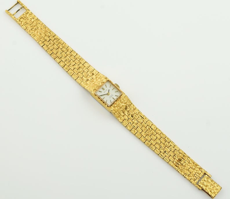 May include: A gold-toned wristwatch with a textured band and rectangular face. The watch features a white dial with black hands and markers. The band is made of small, interconnected textured gold links. The watch is displayed against a white background.