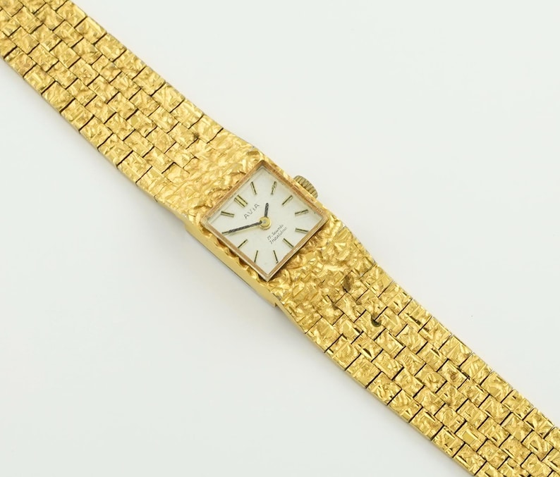 May include: A gold-toned Avia wristwatch with a square face and a textured band. The watch face is white with black numerals and hands. The band has a woven pattern.