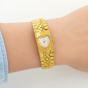 May include: A gold-tone quartz watch featuring a heart-shaped white dial with black hands and markers. The wide, textured metal band displays an engraved leaf pattern above and below the watch face, with a small crown on the right side.