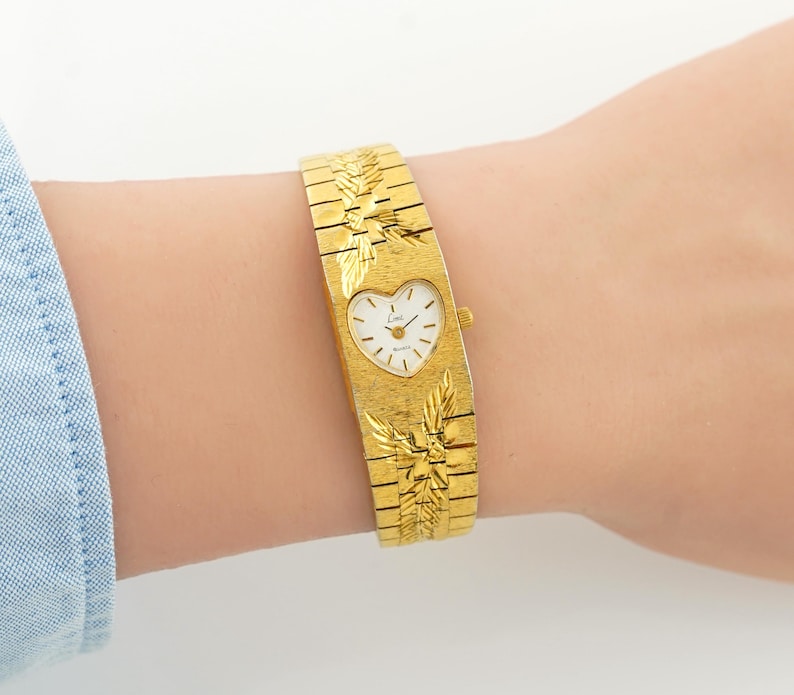May include: A gold-tone quartz watch featuring a heart-shaped white dial with black hands and markers. The wide, textured metal band displays an engraved leaf pattern above and below the watch face, with a small crown on the right side.