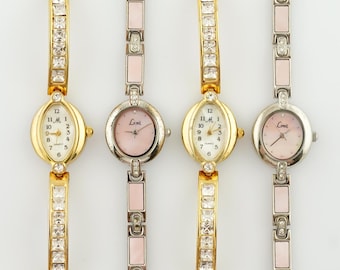 May include: Three gold-tone wristwatches with crystal accents on the band. The watches have oval-shaped faces with white and pink dials. The dials feature black Roman numeral hour markers and black hands. The watches have a silver-tone, gold-tone, and two-tone band.