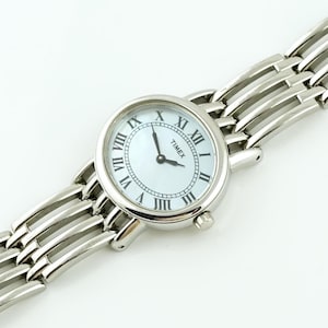 Puede incluir: Un reloj Timex plateado con esfera blanca y números romanos. El reloj tiene una esfera redonda y una correa tipo brazalete. Las manecillas del reloj son negras.